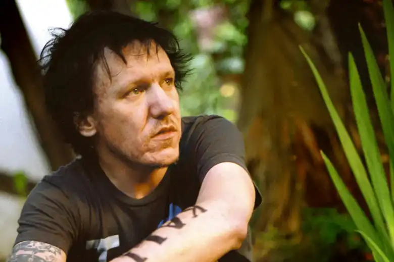 La imagen muestra a Elliott Smith sentado en medio de un jardín mirando a la distancia.