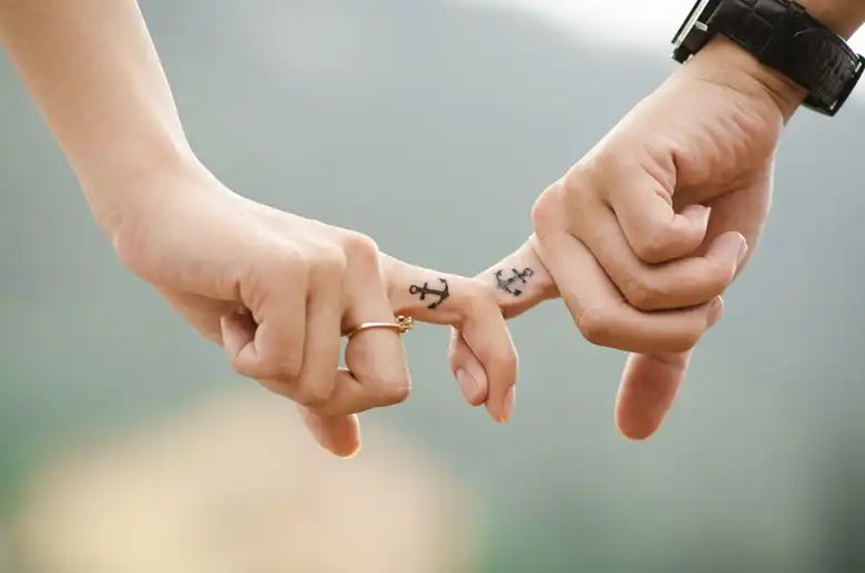 La imagen muestra a una pareja tomada de la mano con sus dedos índices. Ambos portan un tatuaje de un ancla en sus dedos.